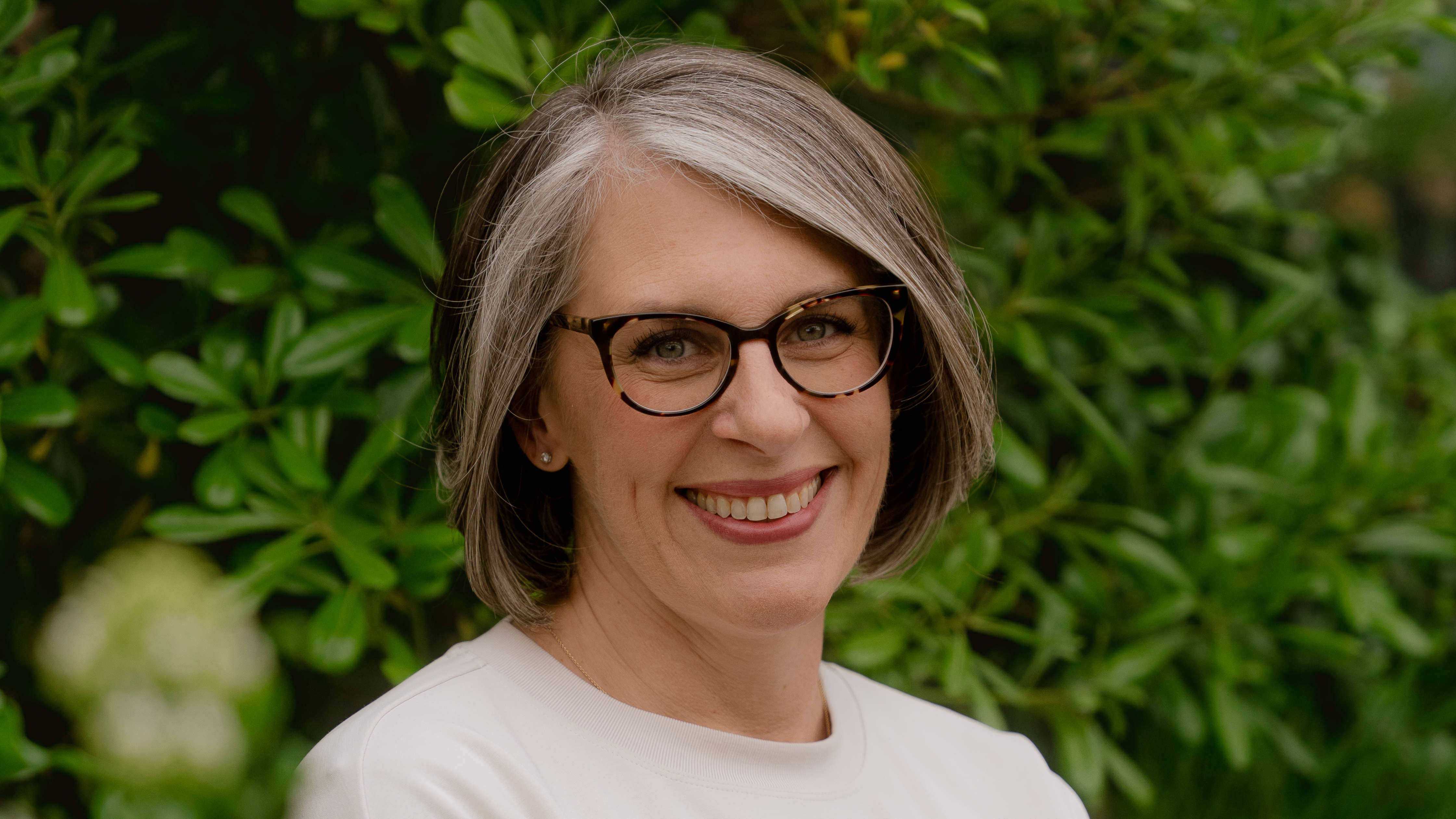 A smiling woman with graying brown hair and glasses poses against a backdrop of lush green leaves.
